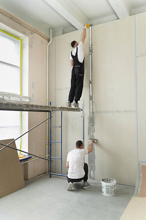 2 men working indoors plastering a wall one standing on a platform